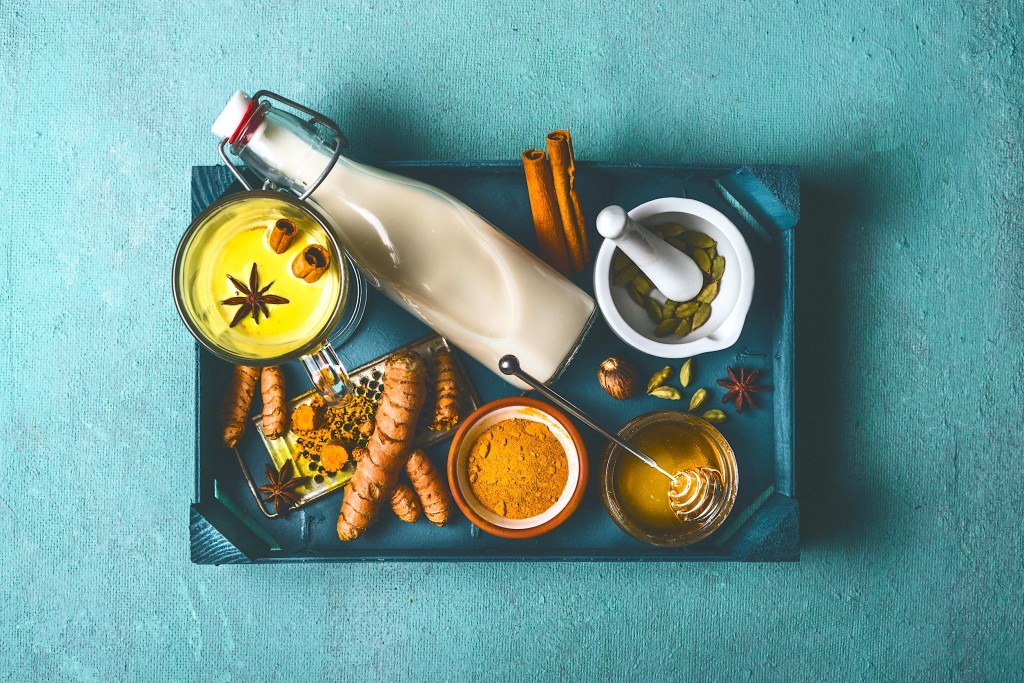 Healthy golden turmeric milk on tray with ingredients: cardamom, turmeric, cinnamon, honey and ginger on blue background , top view, vertical. Immune boosting remedy , detox and dieting concept.
